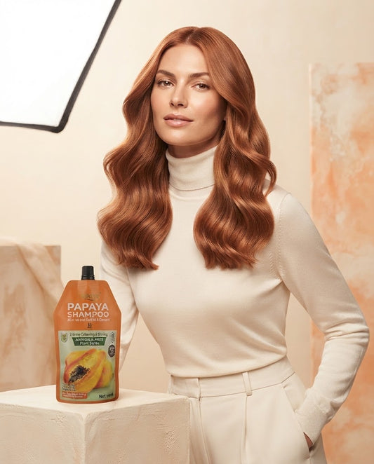 A woman with long, wavy copper-red hair stands indoors wearing a cream turtleneck and cream pants, next to a bottle of Papaya Shampoo on a white pedestal. The background is a warm beige wall with soft lighting and a studio light visible in the upper left.
Papaya Hair Color orange copper result on hair